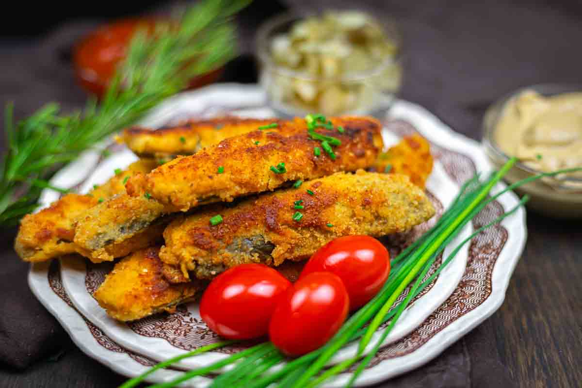 A plate of breaded and fried pickles garnished with fresh chives and served with cherry tomatoes; condiments are in small bowls in the background.