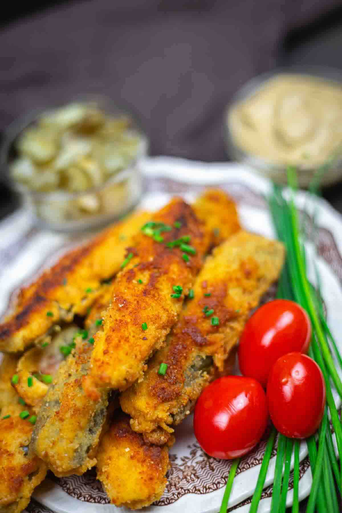 A plate of breaded and fried vegetable sticks garnished with chopped chives, served with cherry tomatoes, fresh chives, and small bowls of pickles and dipping sauce.