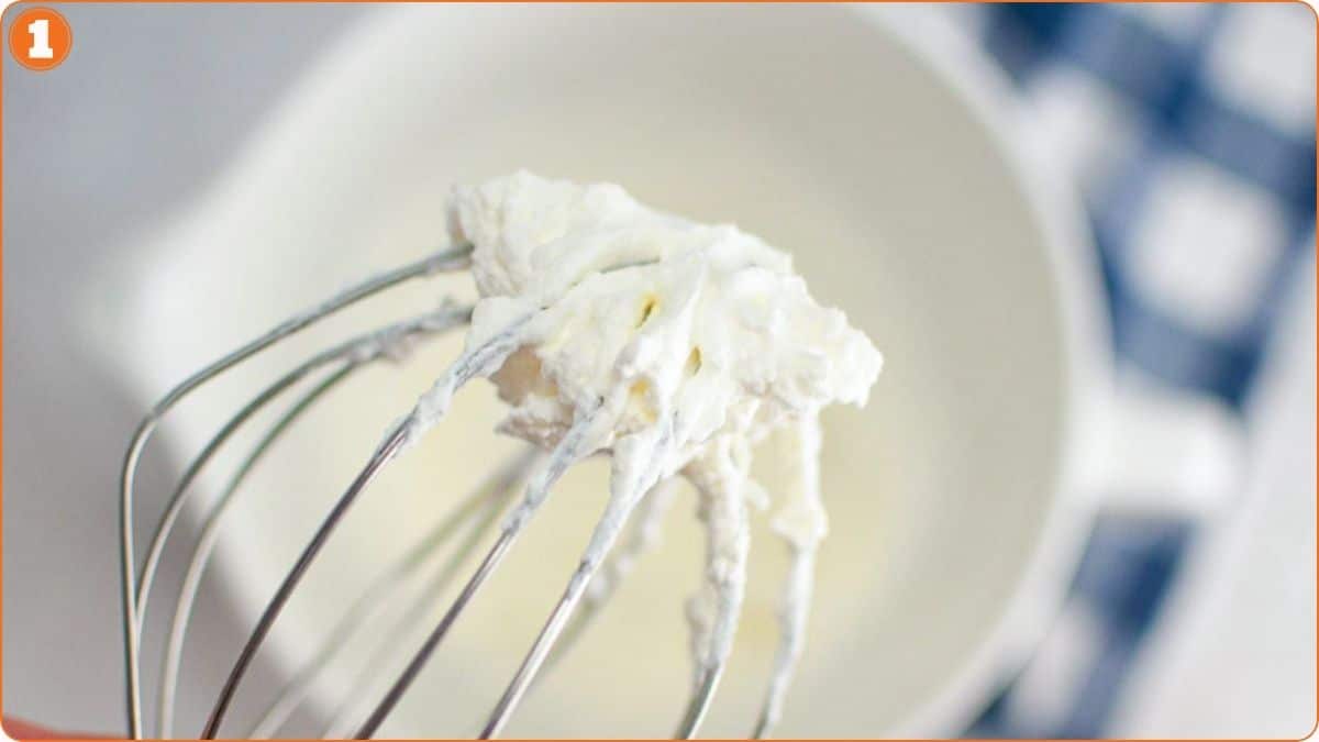 A metal whisk with whipped cream on it is held above a white mixing bowl, with a blue and white checkered cloth in the background.