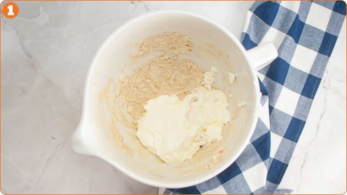 A mixing bowl with partially mixed dough and cream cheese rests on a marble surface next to a blue and white checkered cloth.