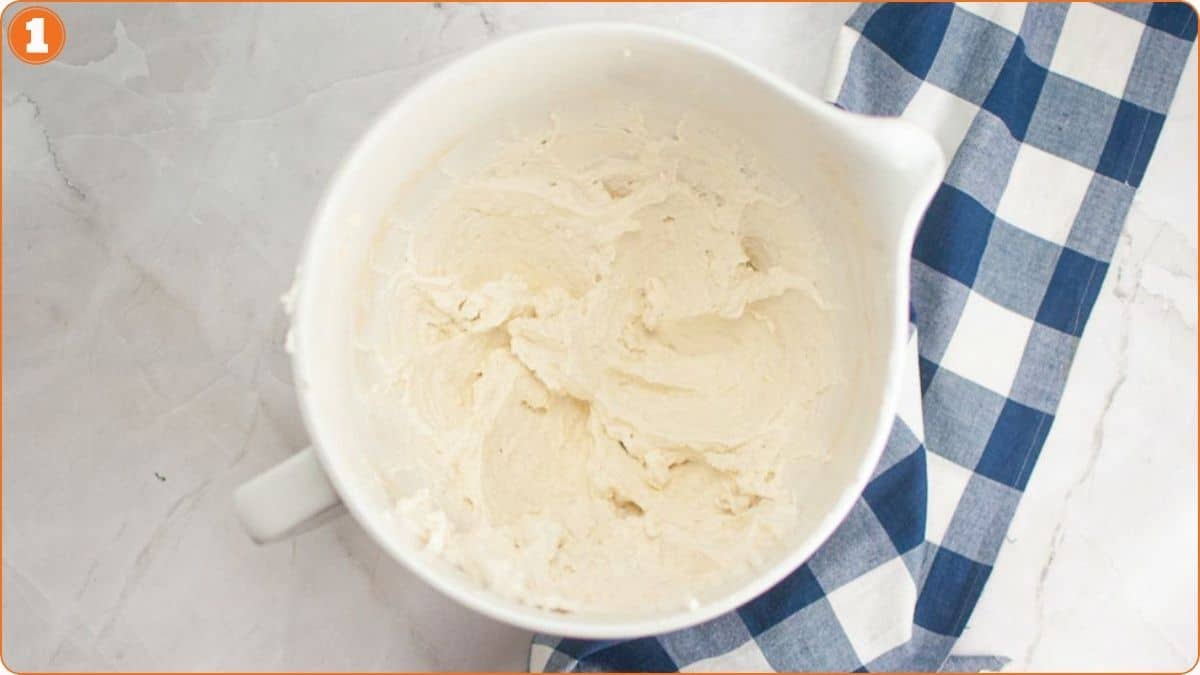 A mixing bowl filled with creamy batter sits on a white marble surface next to a blue and white checkered cloth.