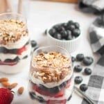 Two glasses filled with layered yogurt, strawberries, blueberries, and granola sit on a table with fresh blueberries, almonds, and a strawberry nearby.