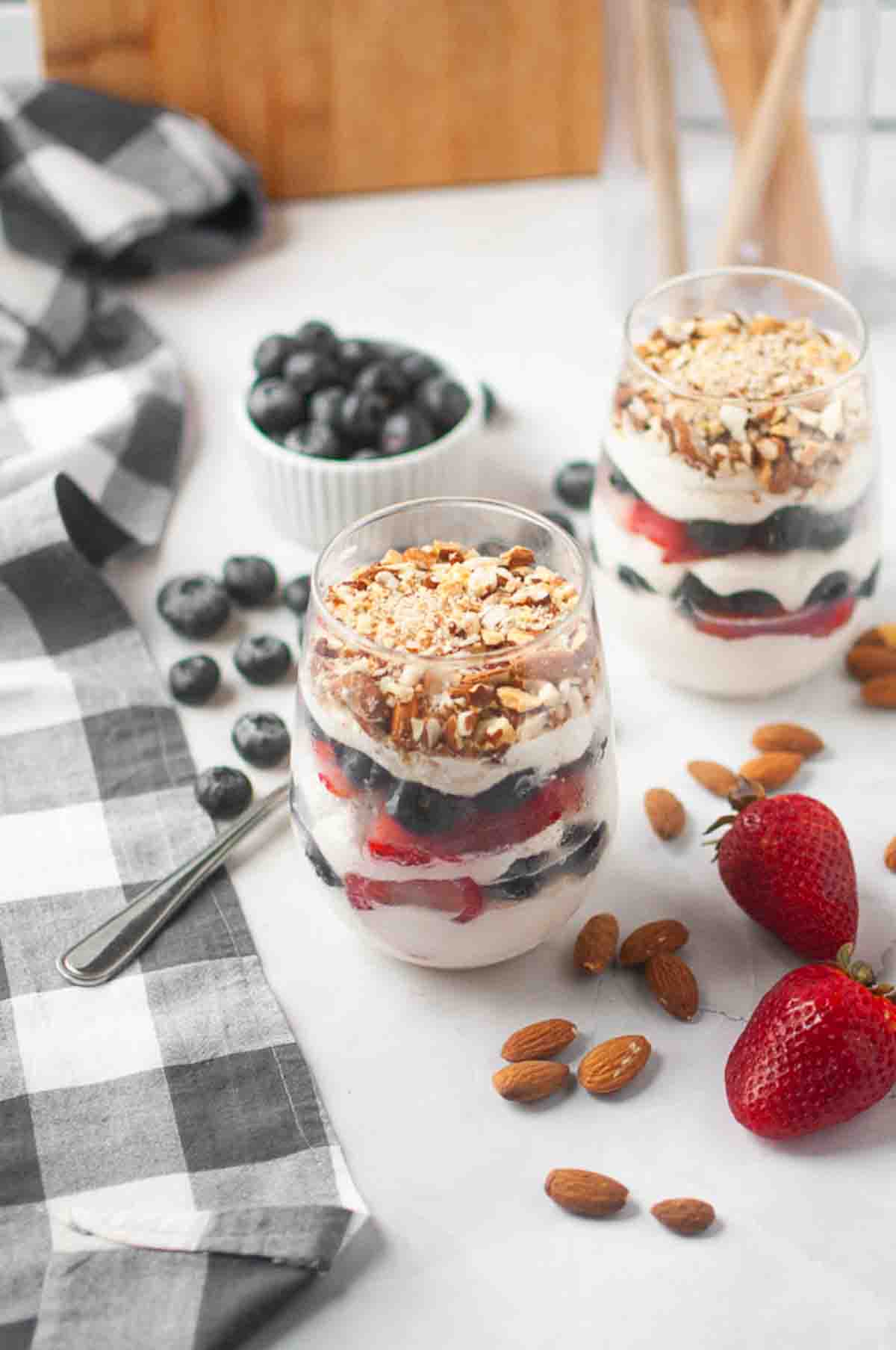 Two glasses filled with layered parfaits of yogurt, strawberries, blueberries, granola, and almonds. Fresh strawberries, blueberries, and almonds are scattered on the table nearby.