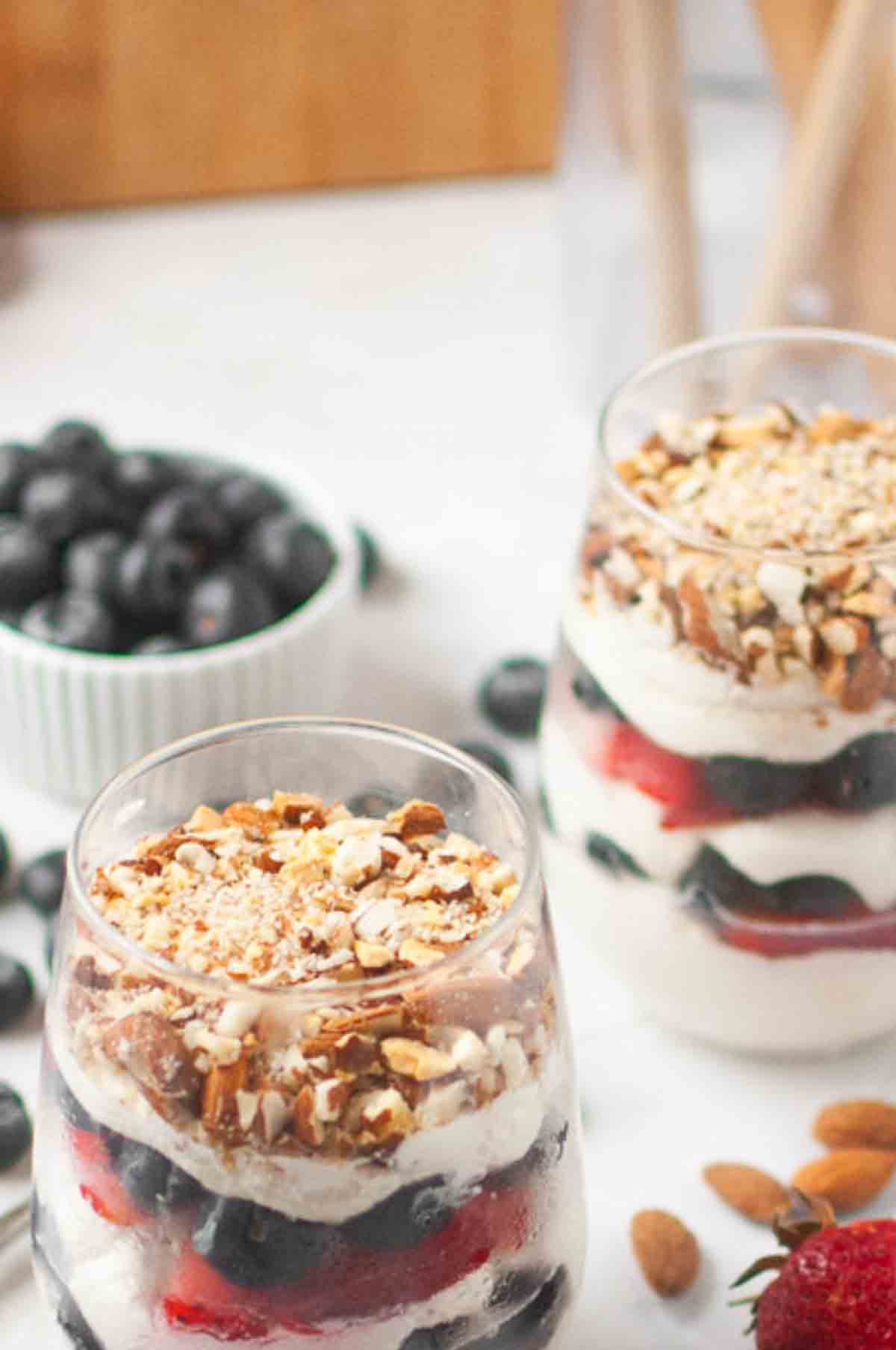Two glasses filled with layered parfaits of yogurt, blueberries, strawberries, and chopped nuts. A small bowl of blueberries and some whole almonds are in the background.