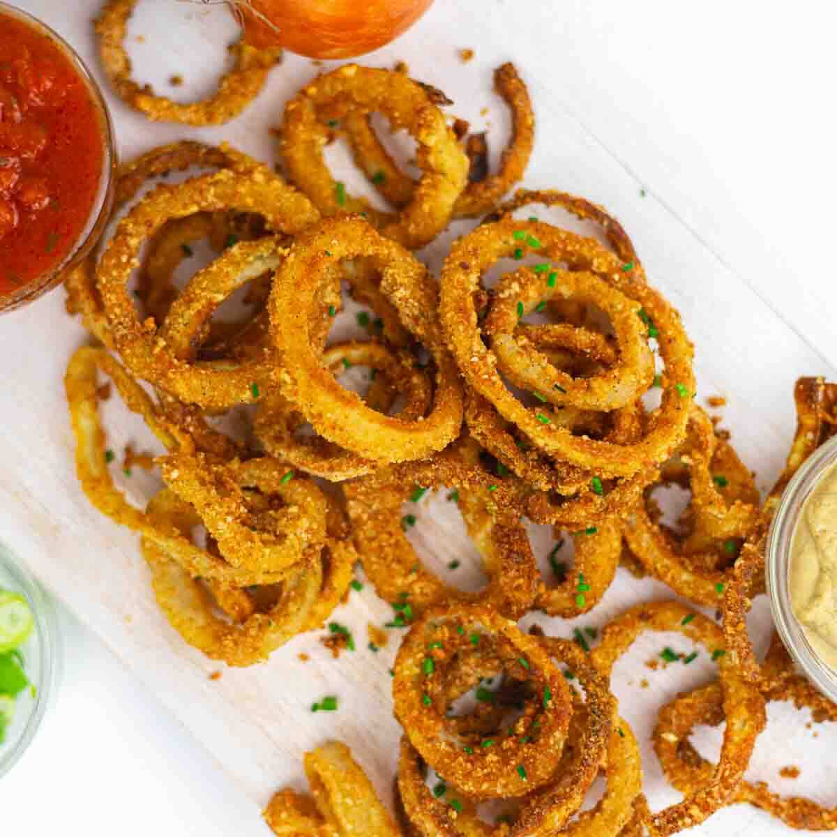 A pile of crispy, golden-brown onion rings garnished with chopped herbs on a white surface, with small bowls of dipping sauces nearby.