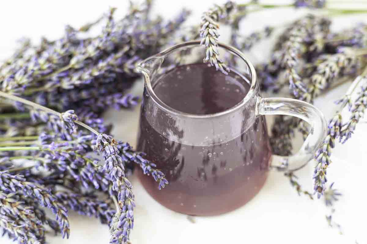 Lavender syrup in glass pitcher surrounded by flowers.