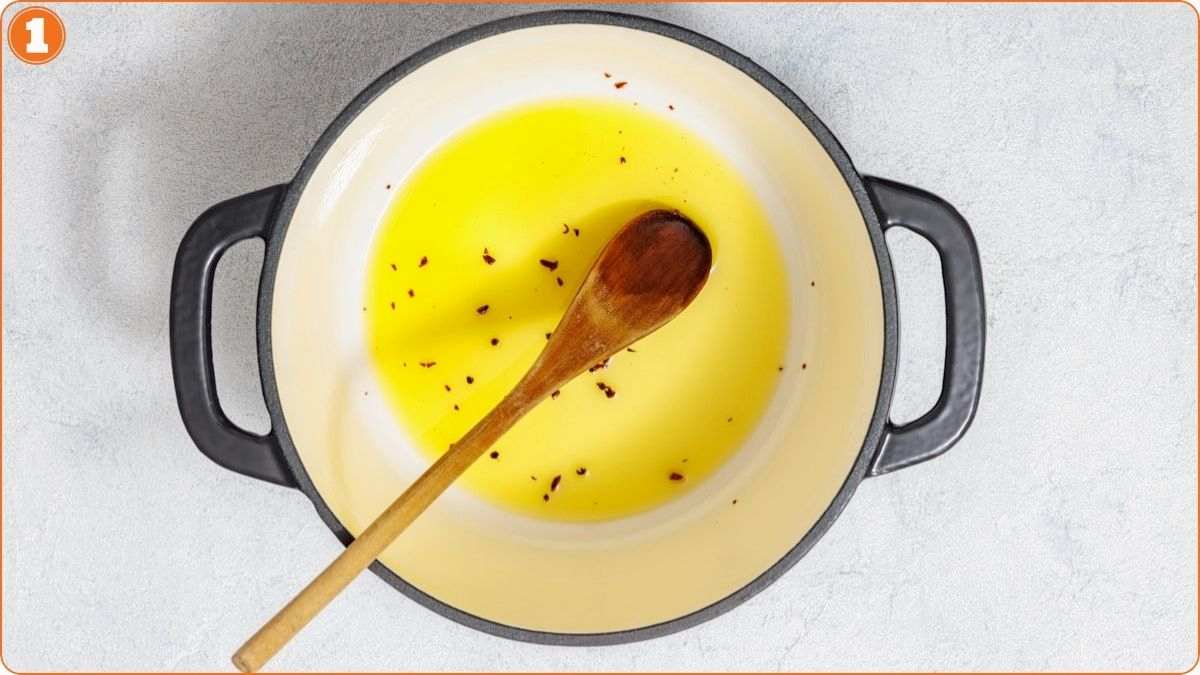 A white pot with two black handles containing yellow oil and red pepper flakes, with a wooden spoon resting inside.