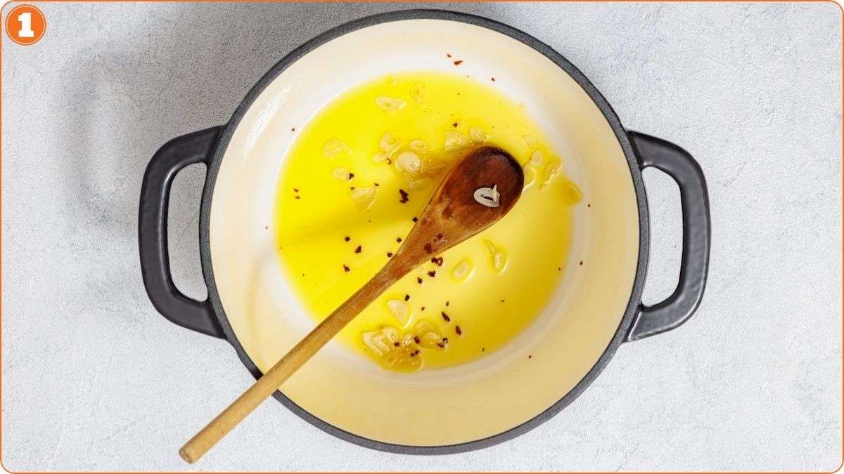 A pot with sliced garlic and red pepper flakes sautéing in oil, stirred with a wooden spoon, on a light-colored surface.