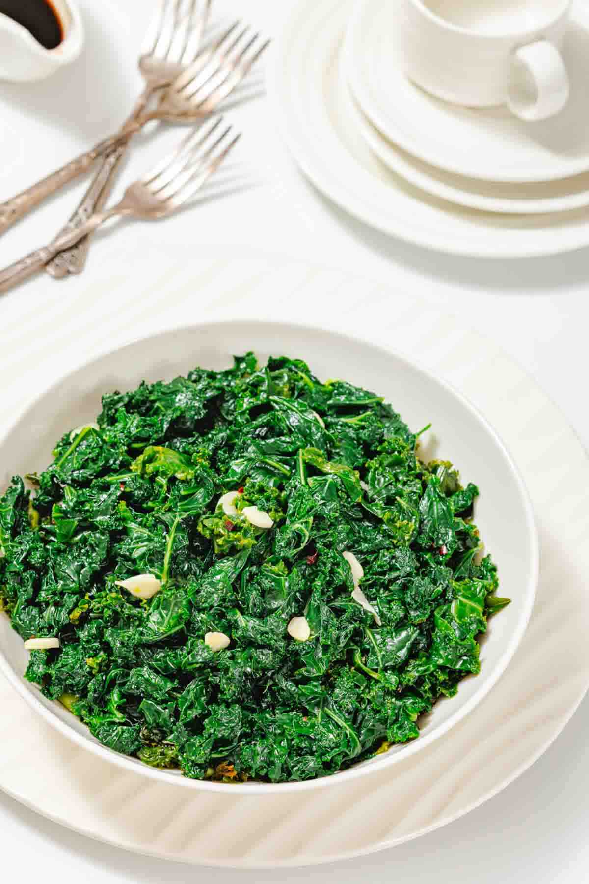 A bowl of sautéed kale with garlic is served on a white plate, set on a table with stacked plates, cups, and forks in the background.