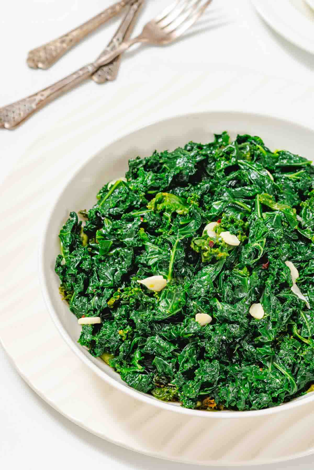 A white bowl filled with sautéed kale and sliced garlic, placed on a round white plate with silver forks in the background.