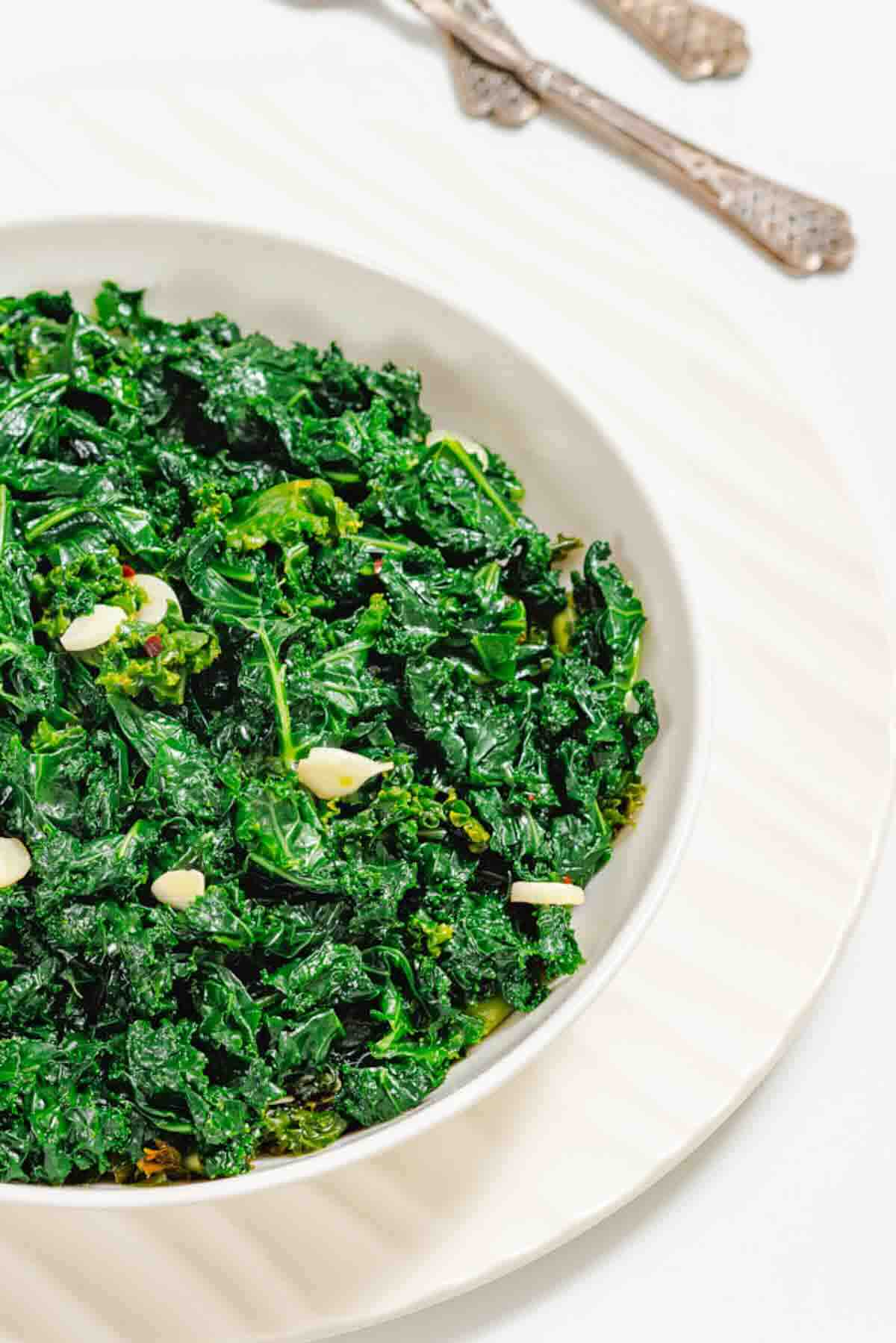 A white bowl filled with sautéed kale and sliced garlic, placed on a white plate with silverware in the background.