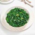 A white bowl filled with cooked, chopped kale garnished with slices of garlic, placed on a white plate with utensils in the background.