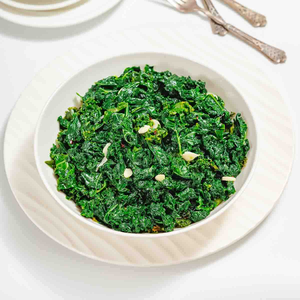 A white bowl filled with cooked, chopped kale garnished with slices of garlic, placed on a white plate with utensils in the background.