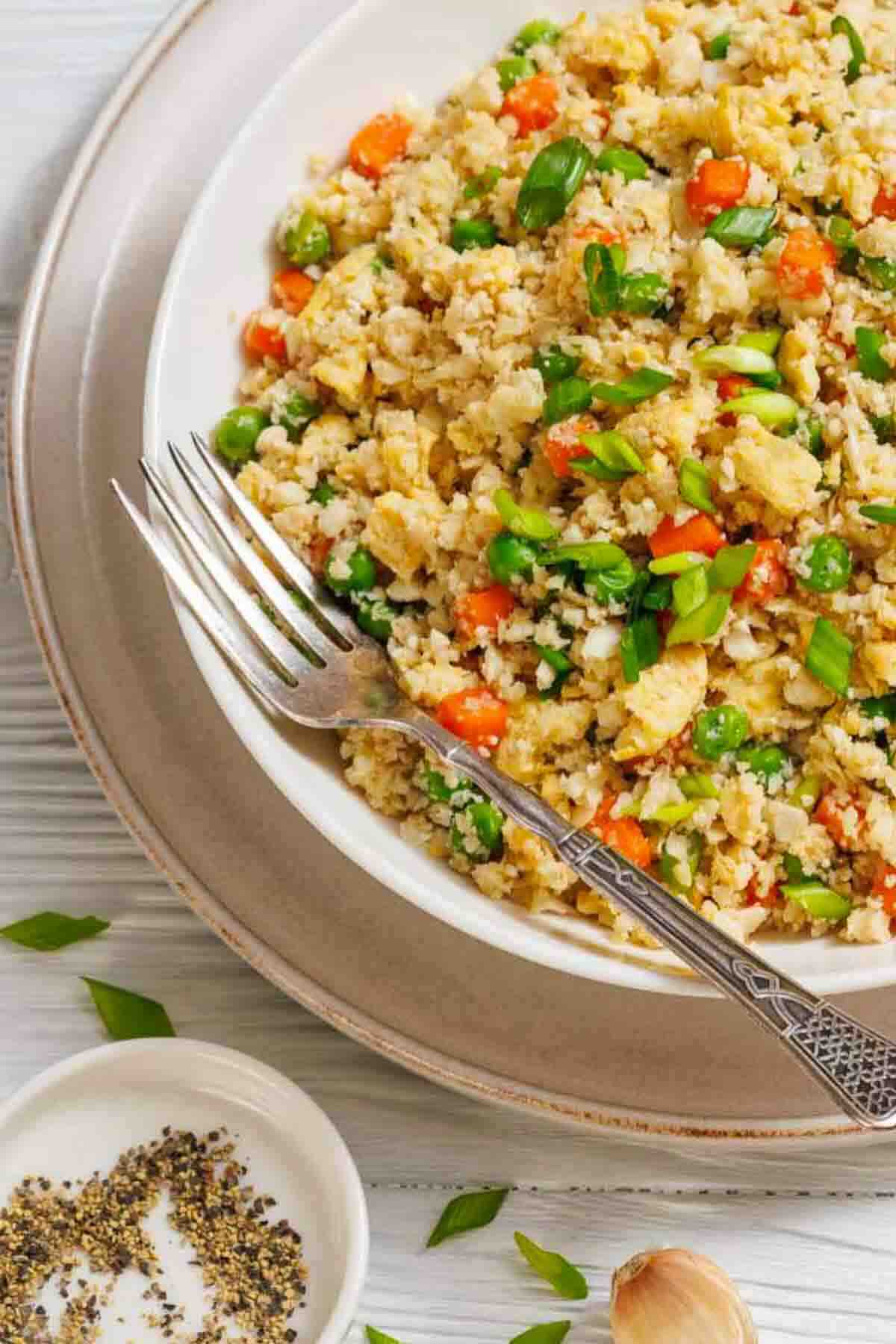 A bowl of vegetable fried rice with peas, carrots, and green onions, served with a fork on a plate. A small dish of cracked black pepper is nearby.
