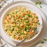 A bowl of cauliflower fried rice with peas, carrots, scrambled eggs, and green onions, placed on a white plate with a fork on the side.