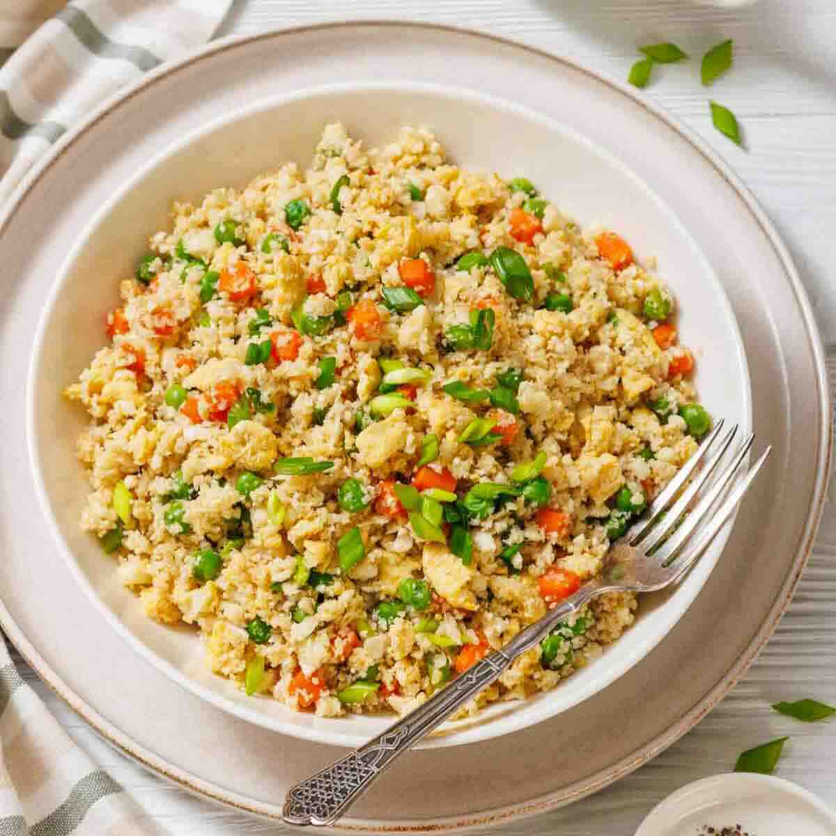 A bowl of cauliflower fried rice with peas, carrots, scrambled eggs, and green onions, placed on a white plate with a fork on the side.