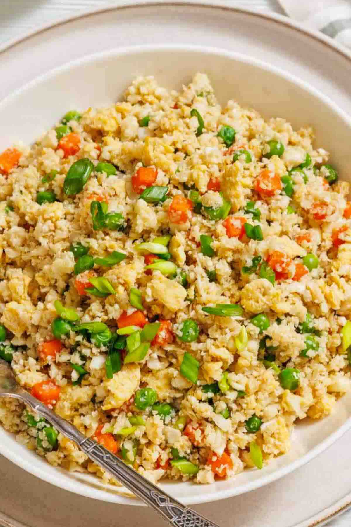 A bowl of cauliflower fried rice with diced carrots, peas, scrambled egg pieces, and chopped green onions, served with a spoon.