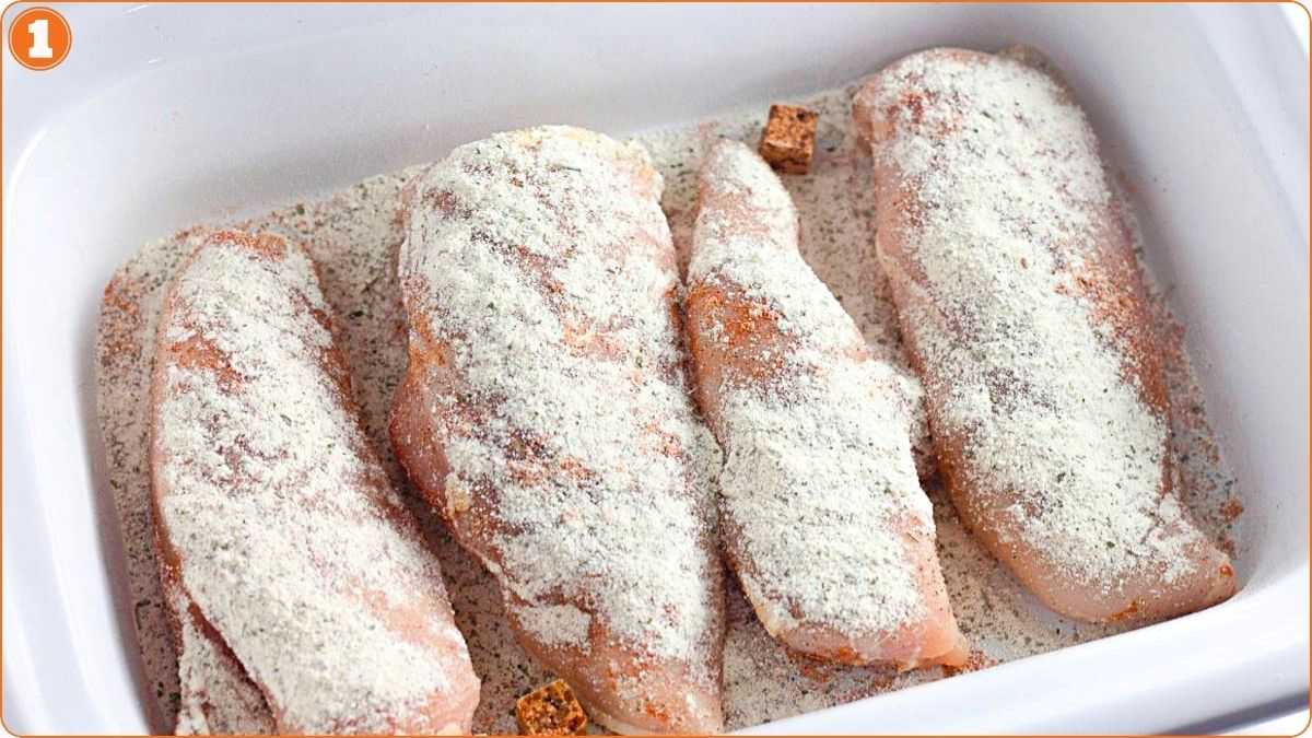Four raw chicken breasts in a white baking dish, coated with a layer of white powdered seasoning and a few brown seasoning cubes.