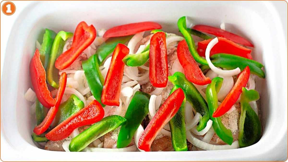 Sliced onions, red bell pepper, and green bell pepper arranged over pieces of raw chicken in a white baking dish.