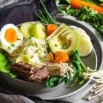 A bowl of ramen noodles topped with sliced boiled egg, avocado, beef, mushrooms, spinach, carrots, and green onions. Fresh vegetables are arranged around the bowl.