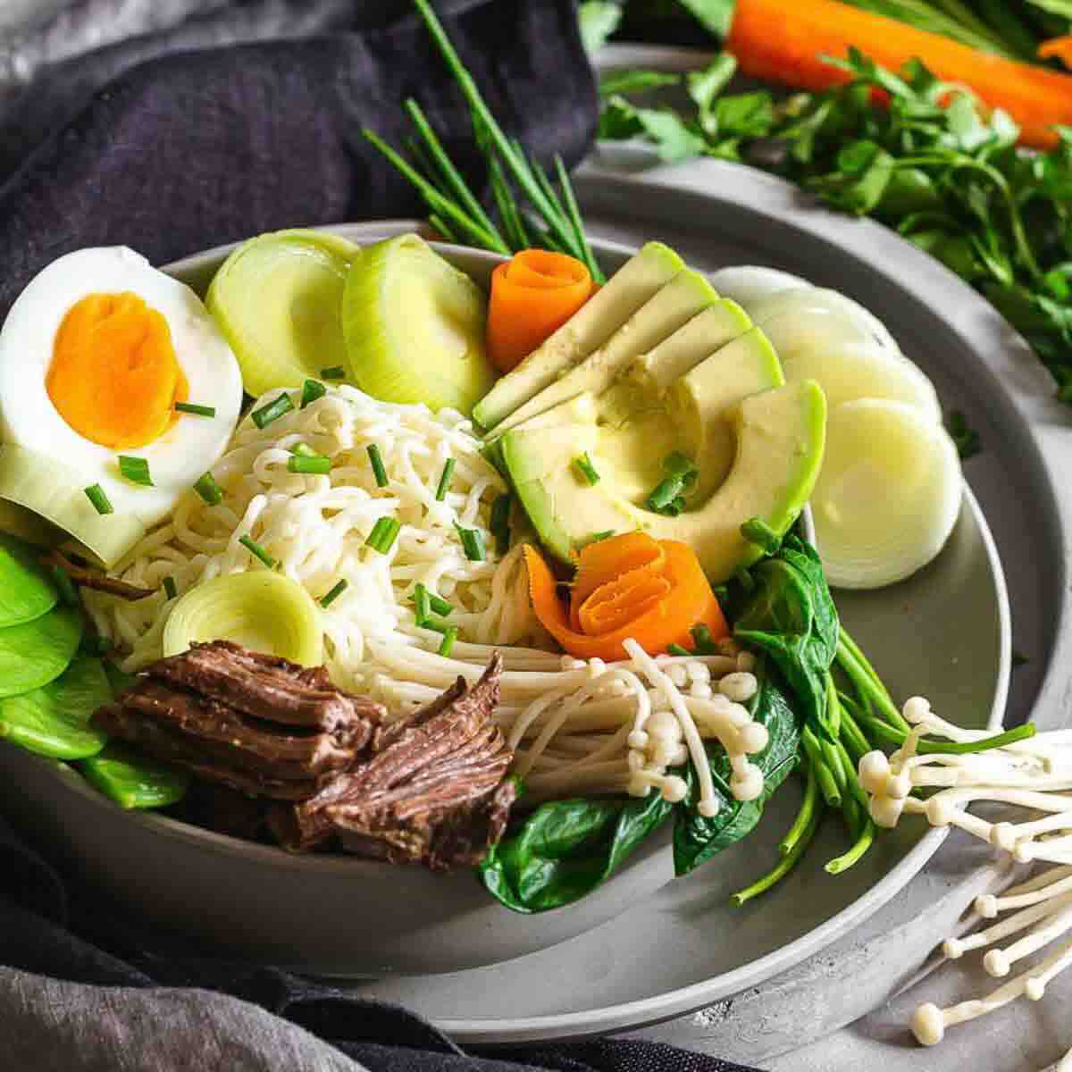 A bowl of ramen noodles topped with sliced boiled egg, avocado, beef, mushrooms, spinach, carrots, and green onions. Fresh vegetables are arranged around the bowl.
