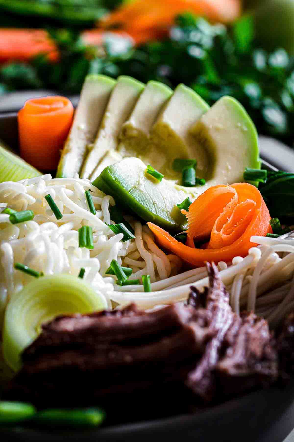 Close-up of a bowl containing noodles, sliced avocado, rolled carrot slices, green onions, and shredded meat, with fresh herbs in the background.