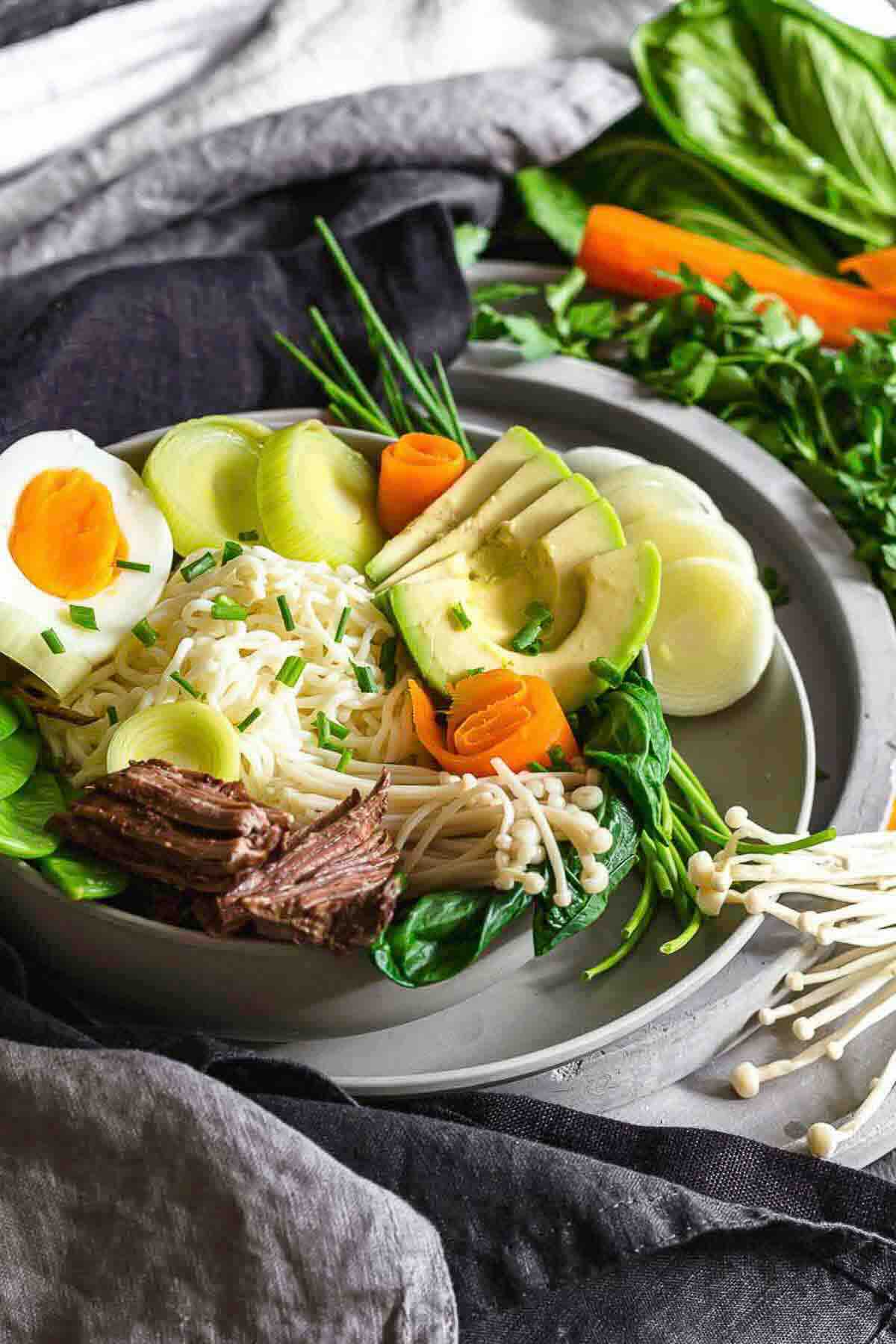 A bowl of ramen noodles topped with sliced avocado, hard-boiled egg, beef, enoki mushrooms, carrots, spinach, and leeks, with fresh vegetables on the side.