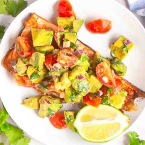 A plate of grilled fish topped with avocado and tomato salsa, garnished with fresh cilantro and served with a wedge of lime.