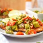 Grilled fish fillet topped with a chunky avocado, tomato, and red onion salsa, served on a white plate with lime slices in the background.