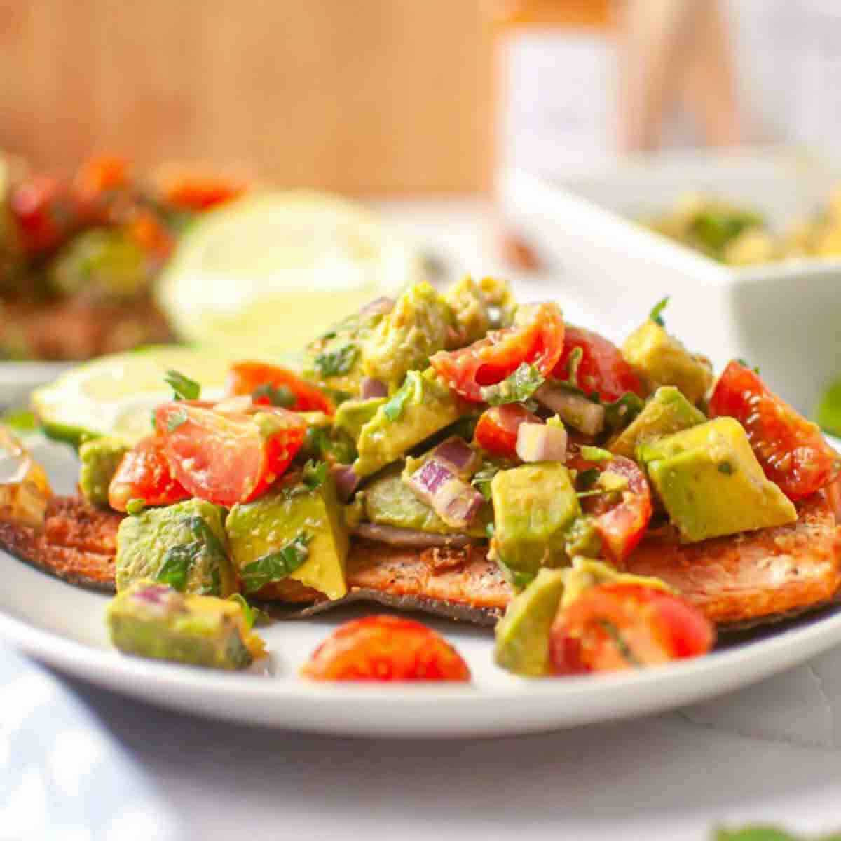 Grilled fish fillet topped with a chunky avocado, tomato, and red onion salsa, served on a white plate with lime slices in the background.