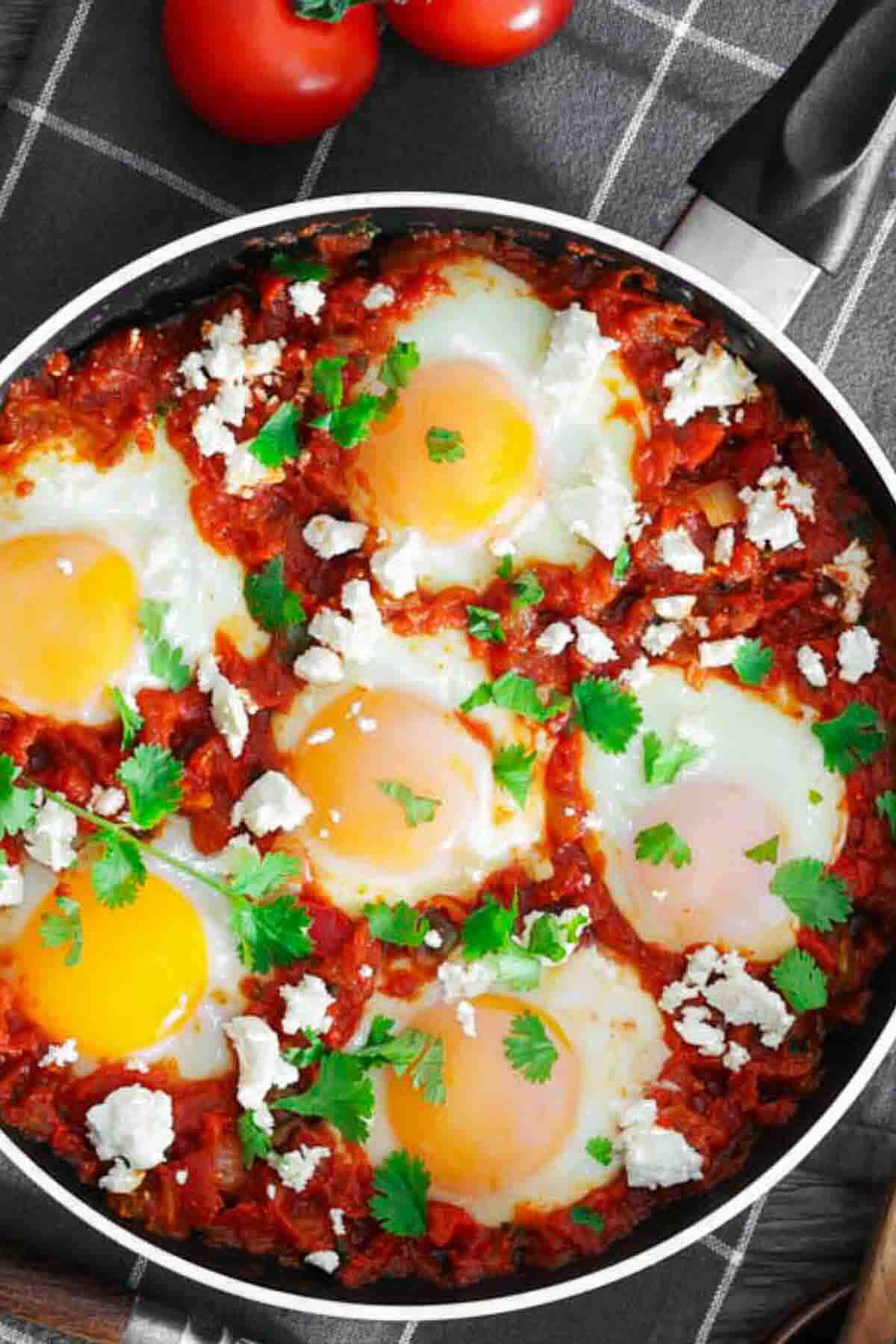 A skillet with poached eggs in a tomato sauce, garnished with herbs and crumbled cheese, sits on a checkered cloth.