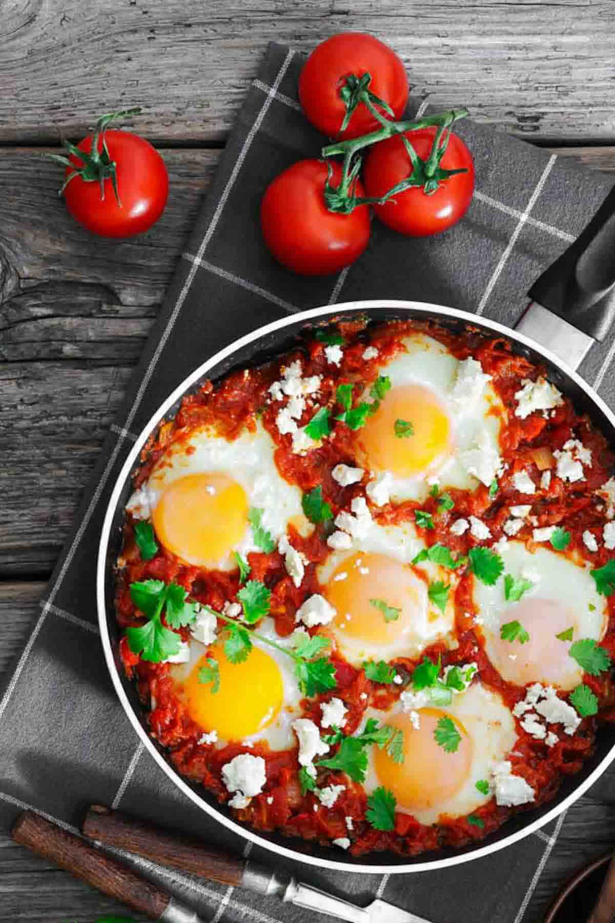 A skillet of shakshuka with poached eggs, tomato sauce, crumbled cheese, and fresh herbs sits on a cloth with three whole tomatoes nearby.
