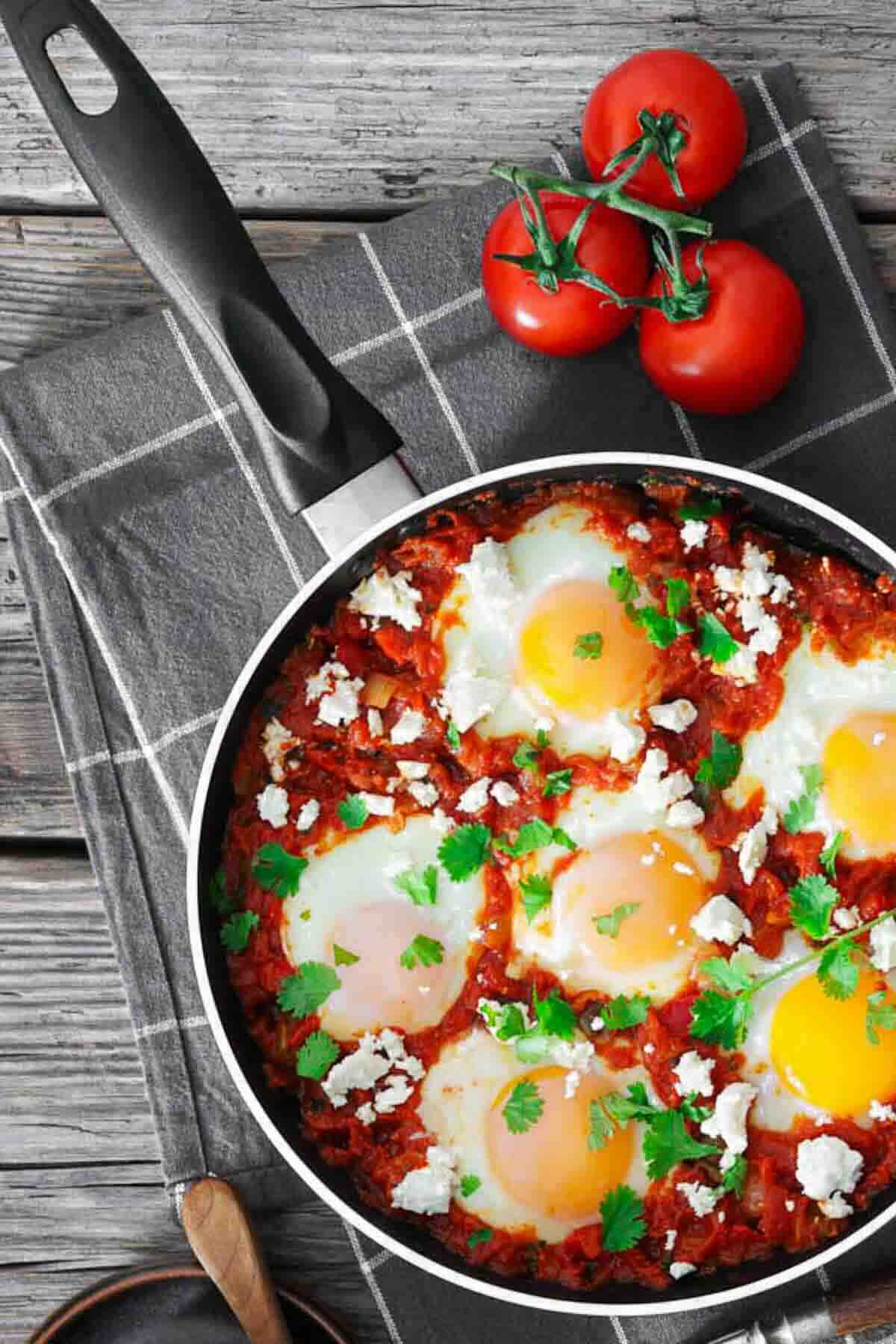 A pan of shakshuka with six eggs, tomato sauce, crumbled cheese, and fresh parsley sits on a checkered cloth next to a vine of tomatoes.