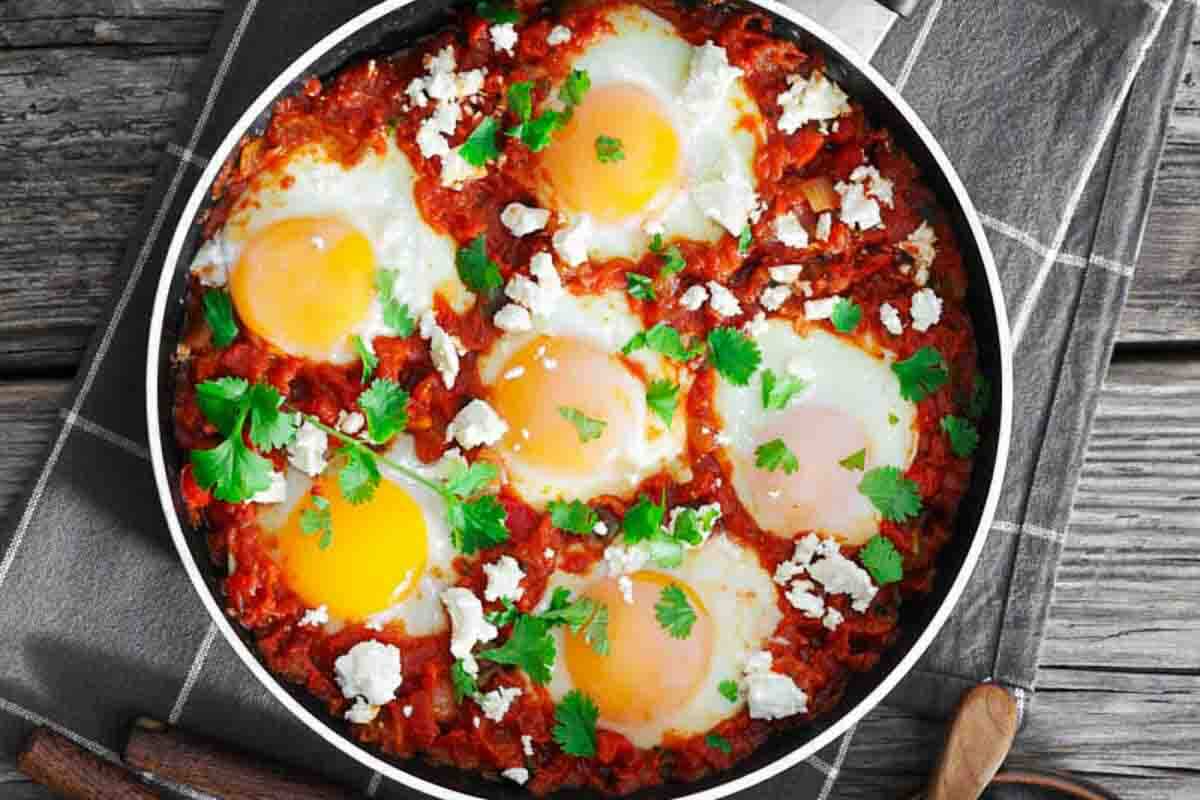 A skillet with six eggs cooked in a tomato-based sauce, garnished with fresh cilantro and crumbled cheese, on a checkered towel atop a wooden surface.