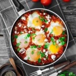 A pan of shakshuka with poached eggs, tomato sauce, herbs, and cheese sits on a checkered cloth next to tomatoes and cutlery.