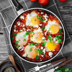 A pan of shakshuka with poached eggs, tomato sauce, herbs, and cheese sits on a checkered cloth next to tomatoes and cutlery.