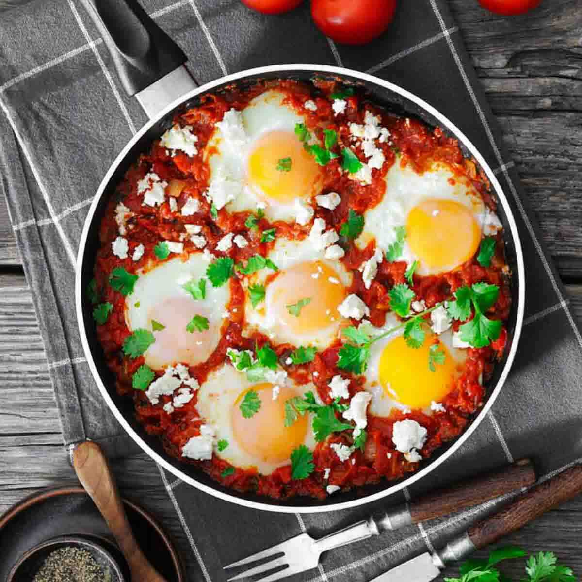 A pan of shakshuka with poached eggs, tomato sauce, herbs, and cheese sits on a checkered cloth next to tomatoes and cutlery.