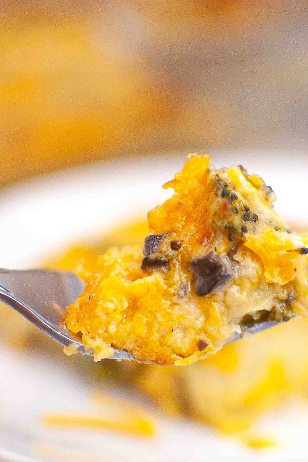 A close-up of a fork holding a bite of cheesy broccoli casserole, with melted cheese and vegetables visible.