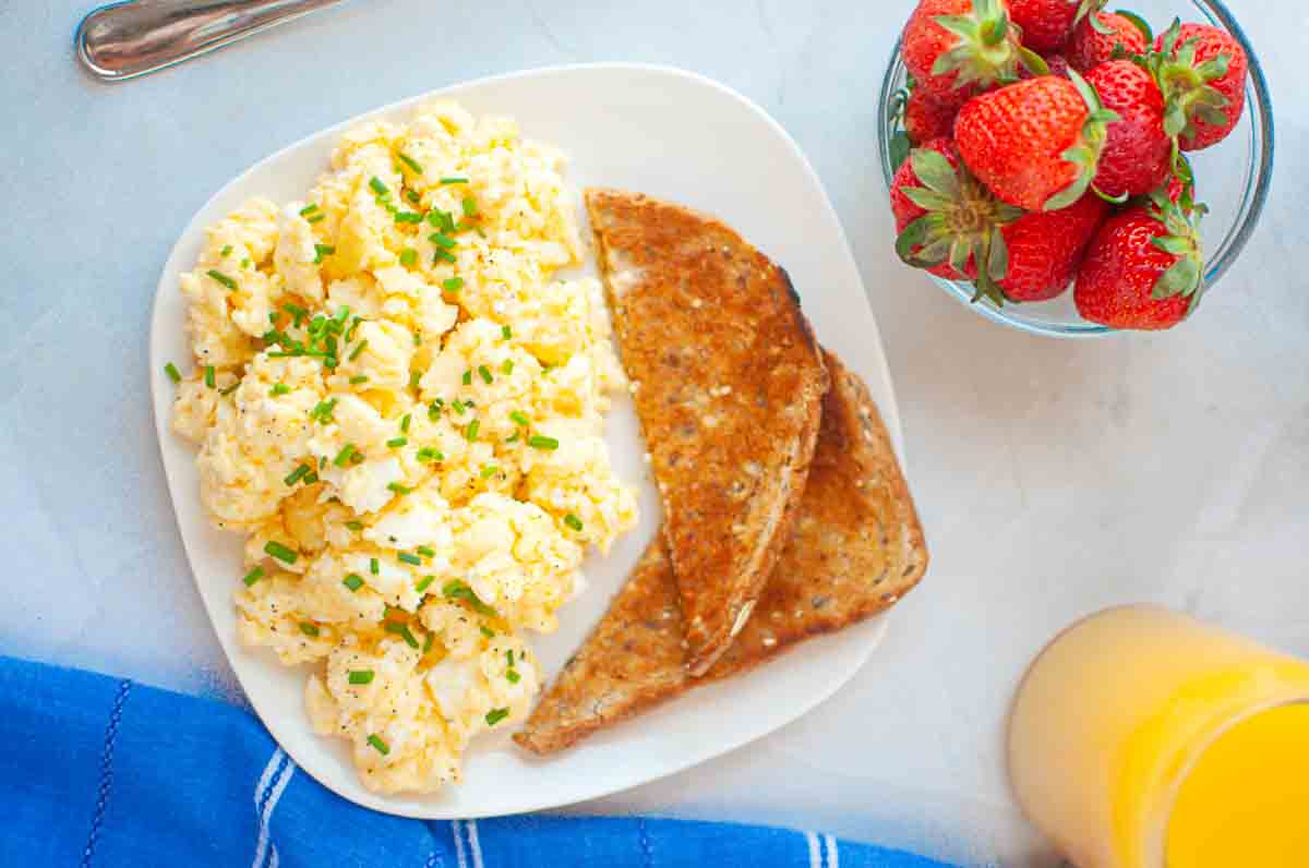 A plate with scrambled eggs topped with chives and two slices of toast, next to a bowl of strawberries and a glass of orange juice.