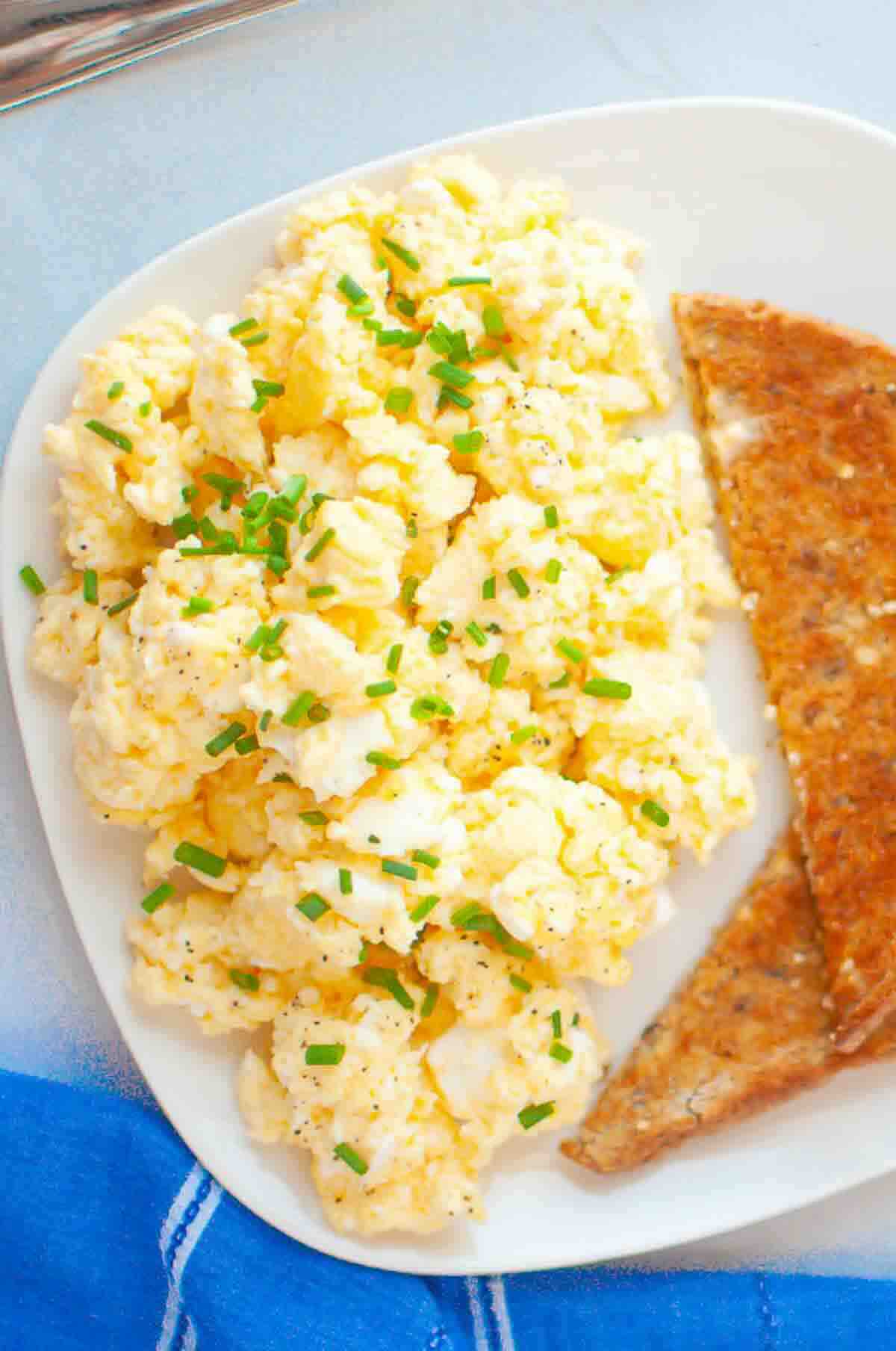 Scrambled eggs topped with chopped chives are served on a white plate with two triangular pieces of toasted brown bread.