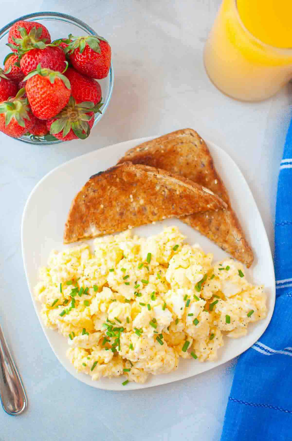 Plate with scrambled eggs garnished with chives, two slices of whole grain toast, a bowl of strawberries, and a glass of orange juice on a table with a blue napkin.