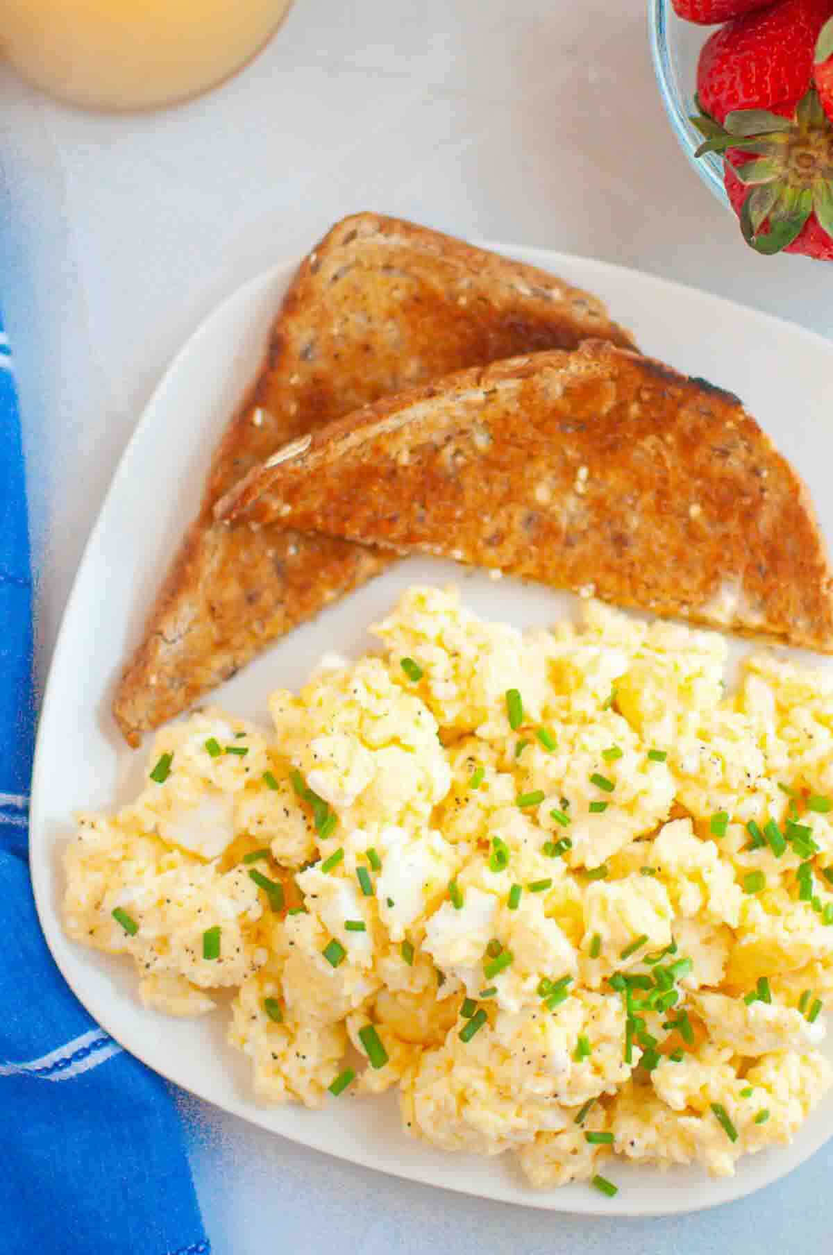 A plate with scrambled eggs topped with chopped chives and two slices of toasted bread, next to a bowl of strawberries.