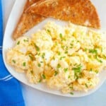 Plate of scrambled eggs topped with chopped chives, served with two pieces of whole grain toast on a white plate next to a blue napkin.