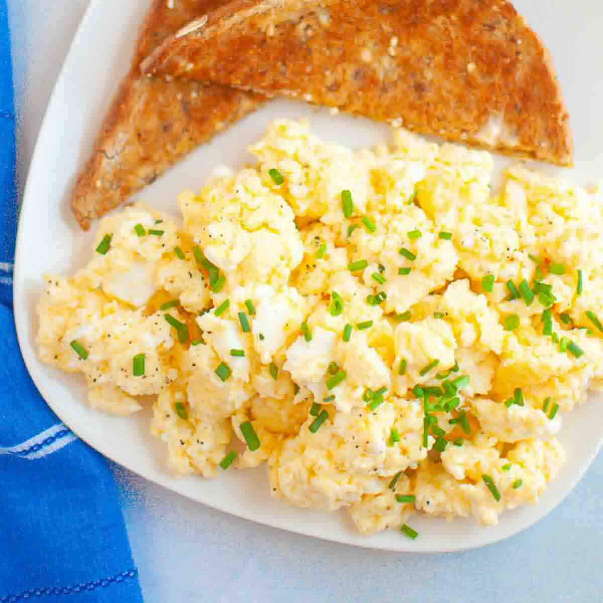 Plate of scrambled eggs topped with chopped chives, served with two pieces of whole grain toast on a white plate next to a blue napkin.