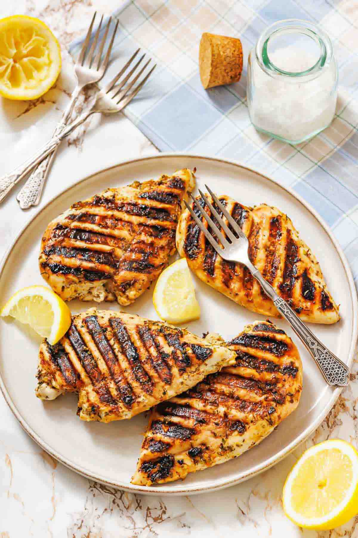 A plate with four grilled chicken breasts, garnished with lemon wedges, sits on a table with forks, a halved lemon, a salt jar, and a cork nearby.