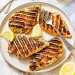 Four grilled chicken breasts with char marks are served on a round plate, garnished with lemon wedges and accompanied by a fork.