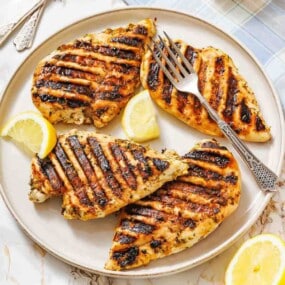 Four grilled chicken breasts with char marks are served on a round plate, garnished with lemon wedges and accompanied by a fork.