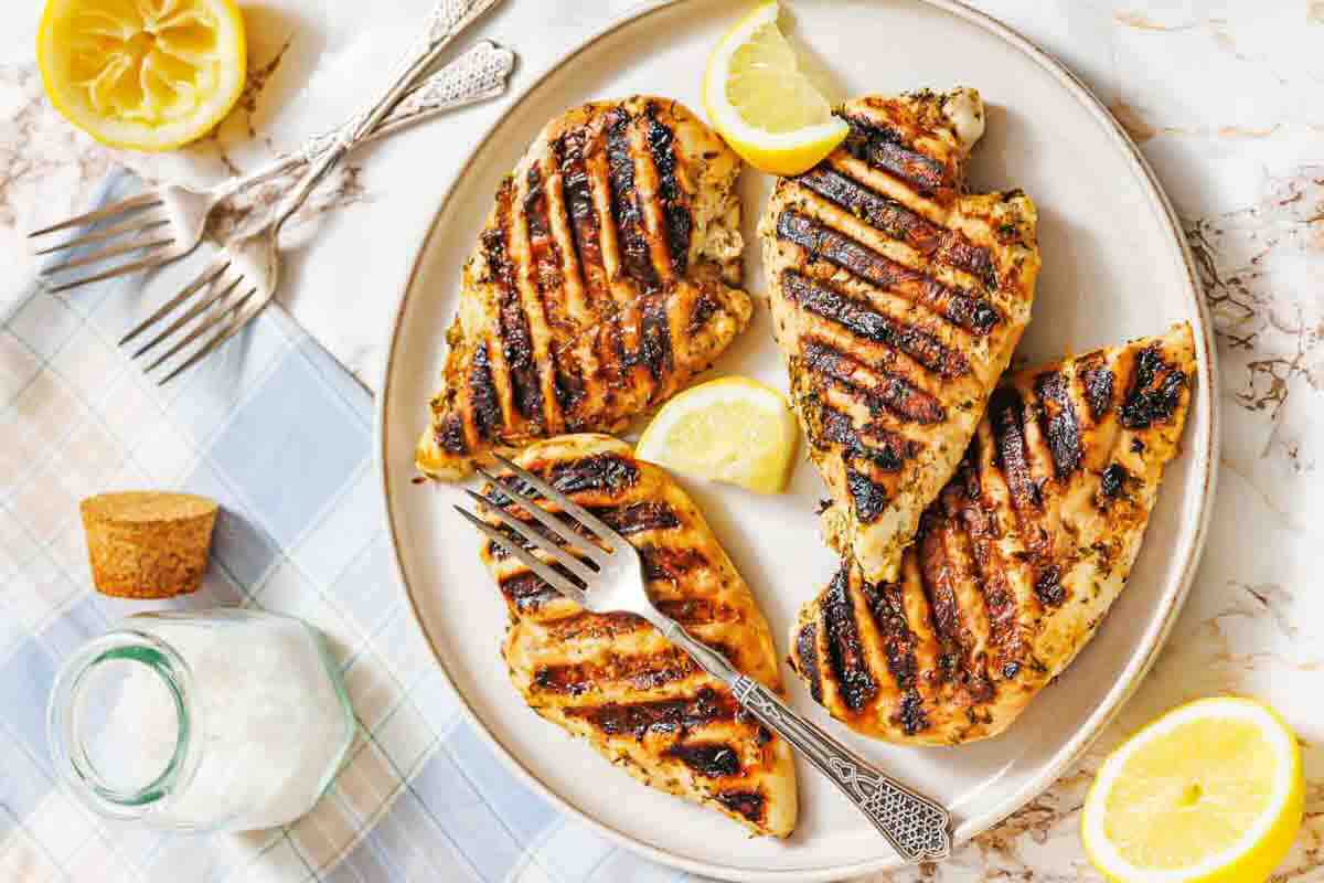 Plate with four grilled chicken breasts garnished with lemon wedges, placed on a table with utensils, a jar of salt, and a halved lemon nearby.