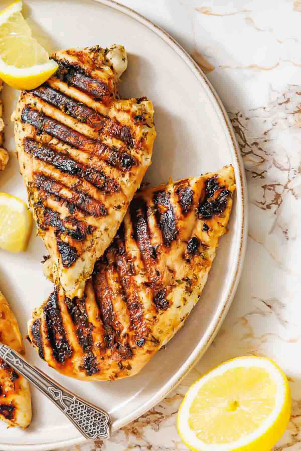 Grilled chicken breasts with visible char marks are served on a round plate with lemon wedges on a marble surface.