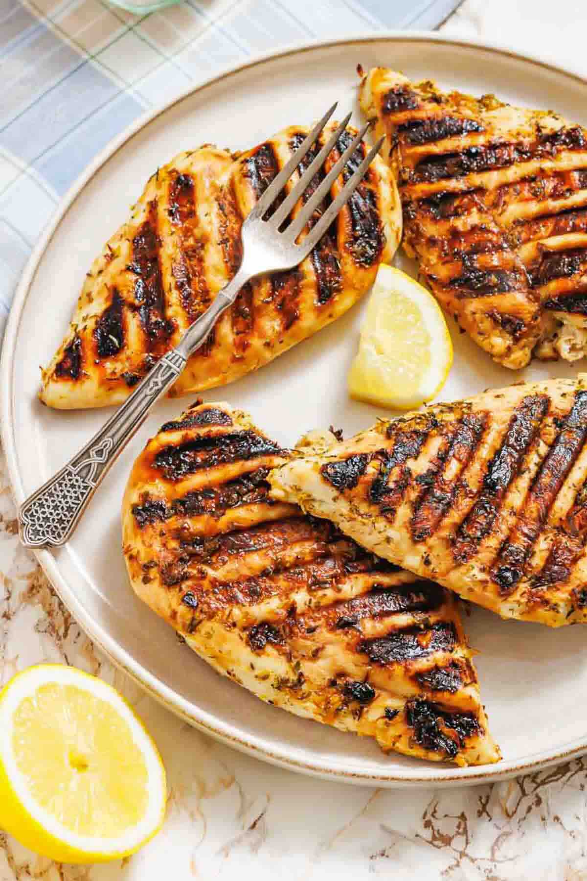 Grilled chicken breasts with char marks on a plate, served with lemon wedges and a fork.
